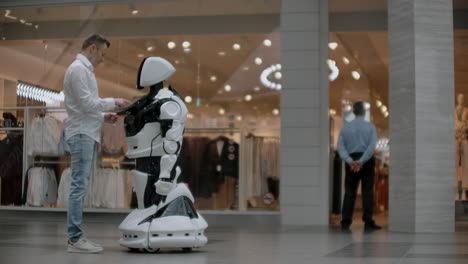 Der-Mann-Im-Einkaufszentrum-Kommuniziert-Mit-Einem-Roboterberater.-Moderner-Laden-Und-Roboterverkäufer.-Roboter-Hilft-Einem-Mann-Im-Einkaufszentrum.